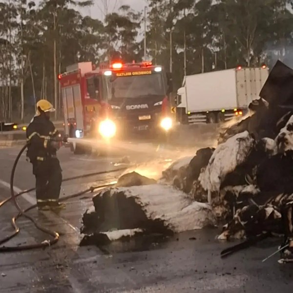 Bombeiro extintando um incêndio em materiais na estrada, com um caminhão de bombeiros ao fundo.