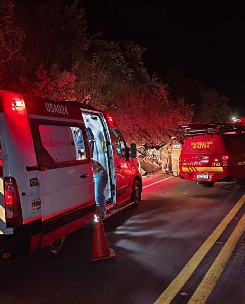 Ambulância e viatura de bombeiros em operação à noite, com luzes vermelhas piscando, próximas a uma área de deslizamento de terra na estrada.