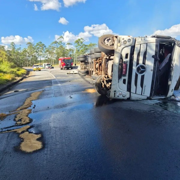 Caminhão tombado na estrada, com óleo derramado, enquanto veículos de emergência estão no local.