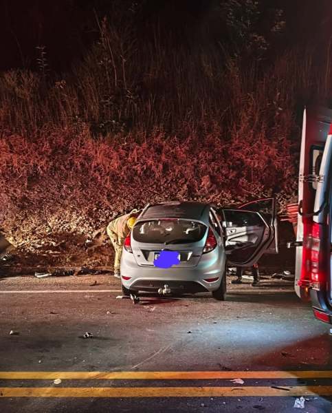 Veículo acidentado na estrada, com uma pessoa inspecionando o carro prata. O ambiente é noturno, iluminado por luzes de emergência.