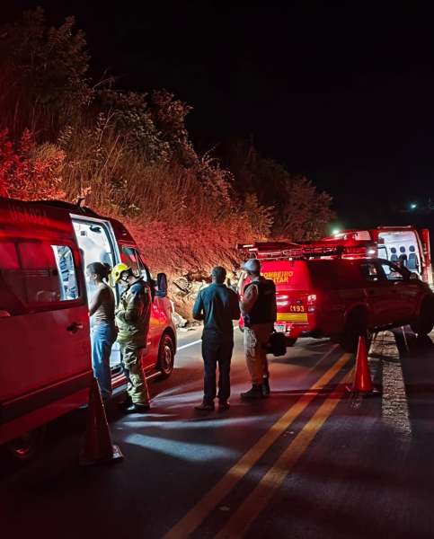 Cena noturna de uma operação de resgate, com veículos de emergência iluminados e pessoas discutindo ao lado de uma estrada.