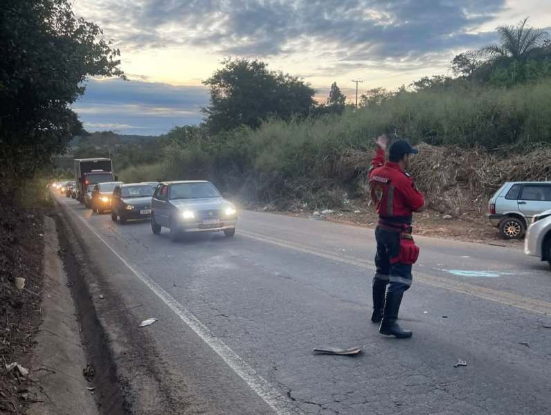 Homem vestindo uniforme vermelho controlando o tráfego em estrada com diversos veículos parados ao longo da pista, ao entardecer.