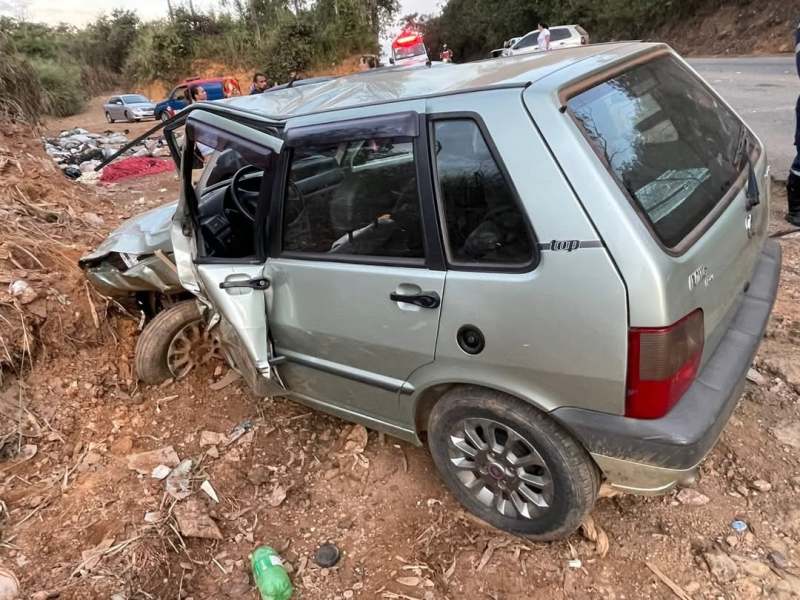 Veículo acidentado em uma área rural, com a porta do motorista danificada e parcialmente aberto, cercado por terra e vegetação.