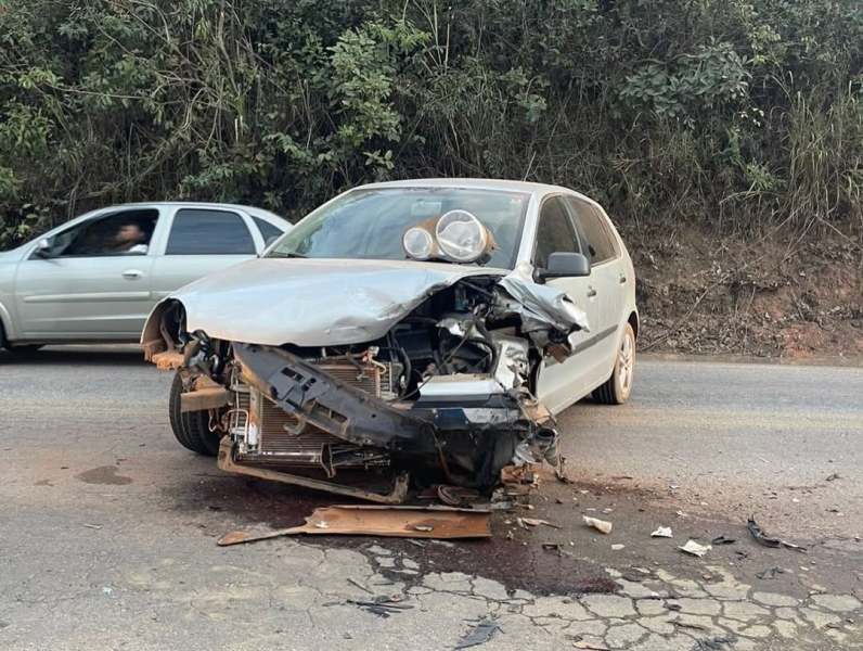 Carro cinza danificado em um acidente, mostrando o motor exposto e destroços na estrada.