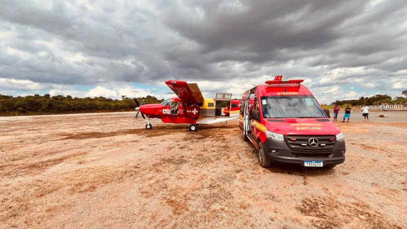 Avião vermelho de combate a incêndio estacionado ao lado de uma ambulância em um campo aberto, sob um céu nublado.