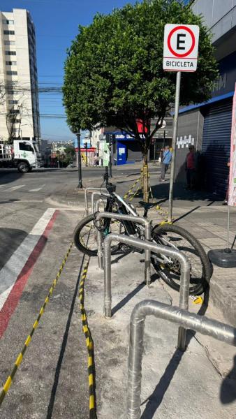 Cicletas presas em suporte na calçada, com sinalização de estacionamento para bicicletas ao fundo e um prédio residencial próximo.