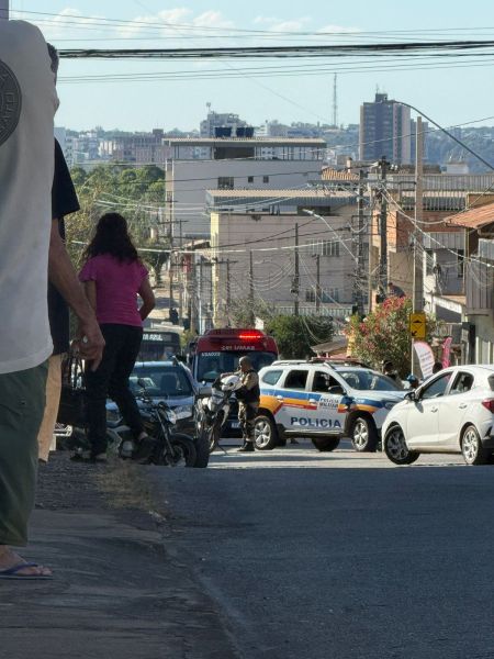Uma rua urbana com carros estacionados e em movimento, incluindo veículos da polícia. Algumas pessoas caminham na calçada, enquanto prédios estão ao fundo.