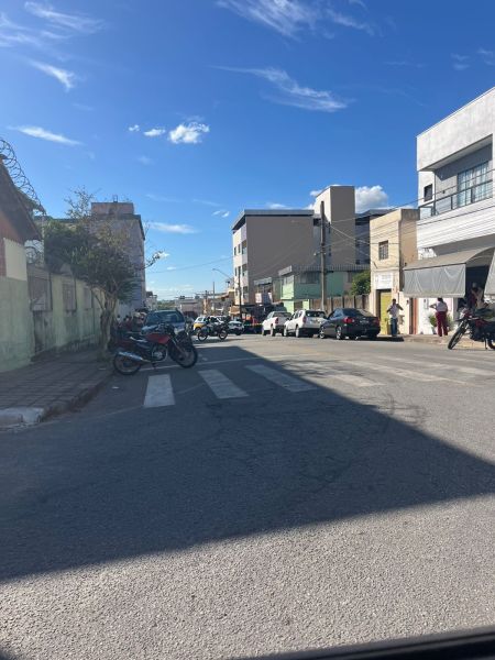 Vista de uma rua com veículos estacionados e andando, prédios ao fundo e céu azul com algumas nuvens.