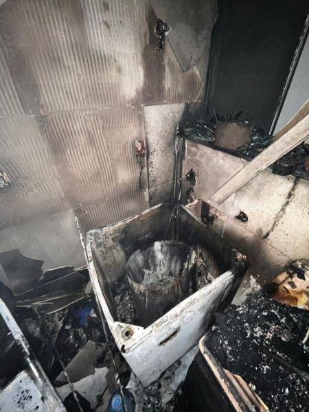 Ambiente interno com um eletrodoméstico queimado, paredes danificadas e resíduos de incêndio visíveis.