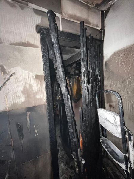 Interior de um espaço afetado por incêndio, com uma porta queimada e paredes danificadas, além de uma cadeira branca visível.