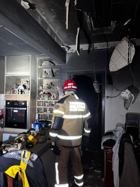 Bombeiro em uma cozinha danificada por incêndio, com fumaça visível e danos estruturais no teto.
