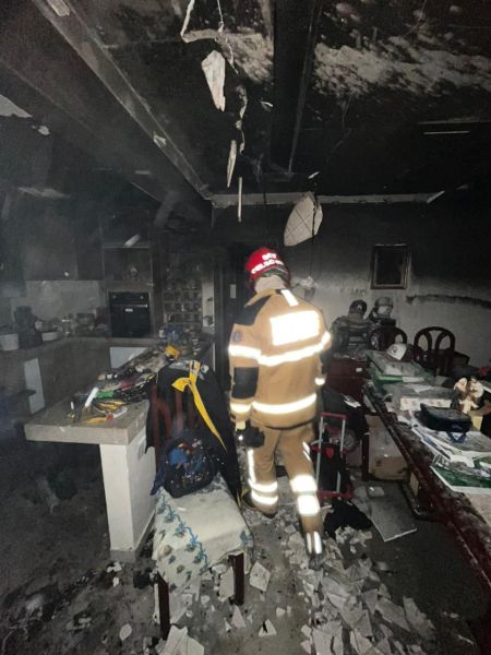 Bombeiro em traje de proteção caminhando em um ambiente danificado por incêndio, com fumaça e escombros visíveis.