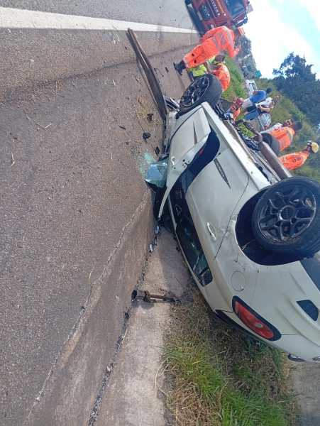 Veículo branco capotado na estrada, com destroços ao seu redor e pessoas uniformizadas prestando assistência.