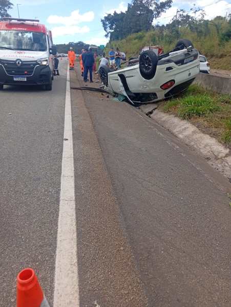 Carro capotado na beira da estrada com pessoas ao redor prestando assistência.