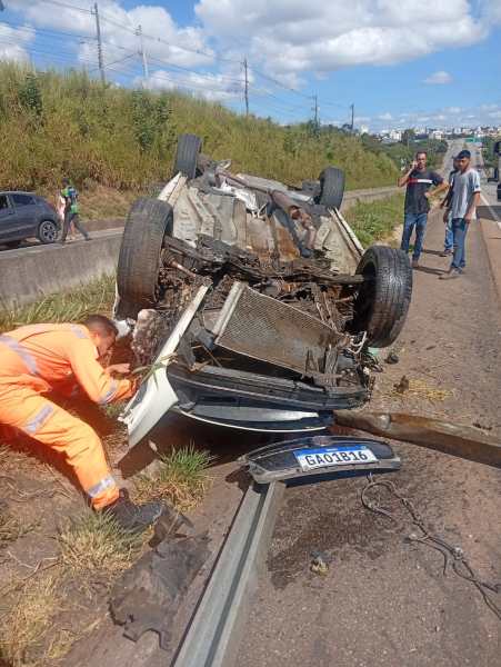 Veículo capotado na estrada, com danos visíveis. Um homem em uniforme laranja está inspecionando o carro, enquanto outras pessoas observam ao fundo.