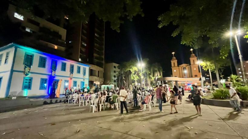 Praça iluminada à noite com pessoas sentadas em cadeiras brancas, um prédio histórico à esquerda e uma igreja ao fundo.