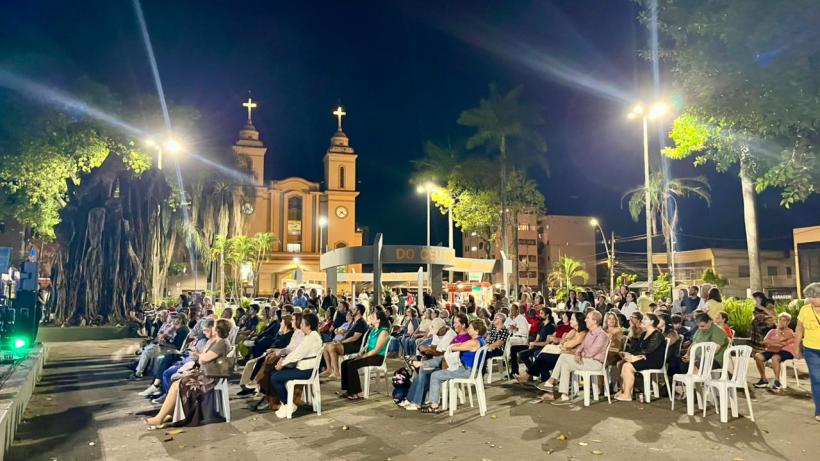Pessoas sentadas em cadeiras plásticas em um evento noturno, com uma igreja ao fundo iluminada por luzes.