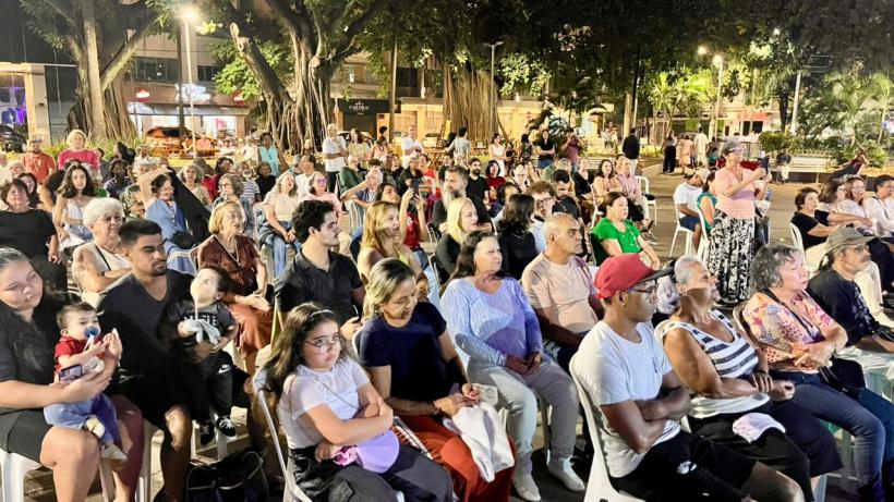 Público assistindo a um evento ao ar livre, com uma multidão de pessoas sentadas em cadeiras, mesclando jovens e idosos em um ambiente iluminado à noite.