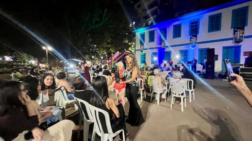 Evento noturno com pessoas sentadas em cadeiras plásticas, várias mulheres conversando e se movimentando em frente a um prédio iluminado em azul.