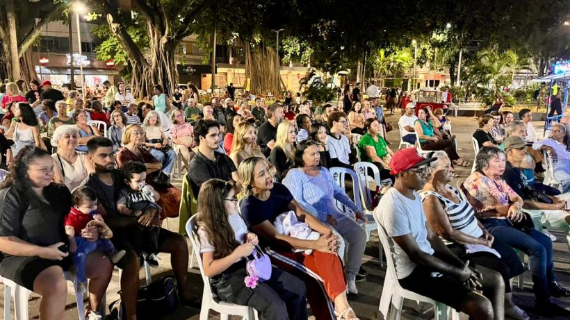 Um grande público sentado em cadeiras plásticas, assistindo a um evento ao ar livre, rodeado por árvores e iluminação noturna.
