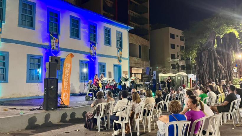 Apresentação musical ao ar livre em praça pública, com um palco iluminado em uma noite clara. Um grupo de músicos toca enquanto o público, sentado em cadeiras brancas, assiste. Ao fundo, um edifício histórico com janelas azuis é iluminado em azul.