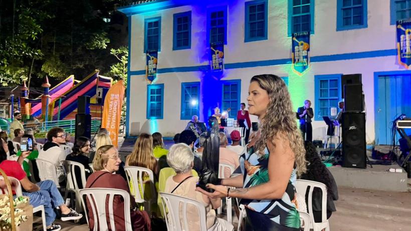 Evento cultural ao ar livre com artistas se apresentando em um palco, enquanto uma plateia sentada participa. Edifício histórico com iluminação colorida ao fundo e brinquedos infantis ao lado.