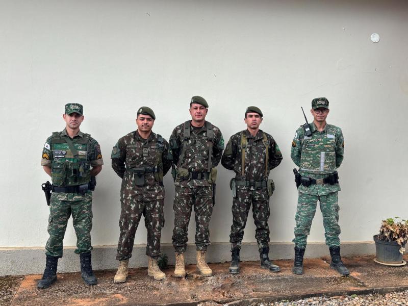 Cinco soldados em pé, vestindo uniformes militares, alinhados em frente a uma parede clara.