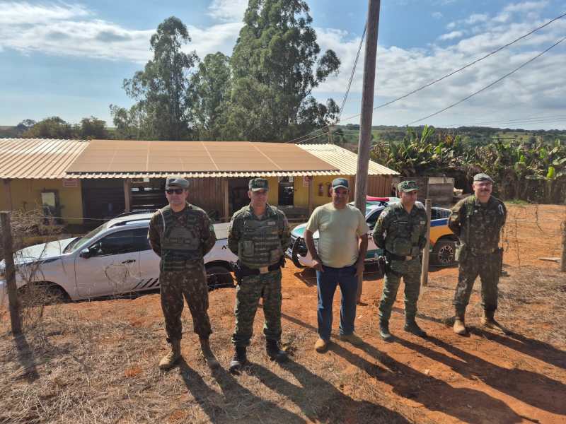 Cinco homens em frente a uma casa amarela, incluindo membros das forças armadas e um civil, com veículos estacionados ao fundo.