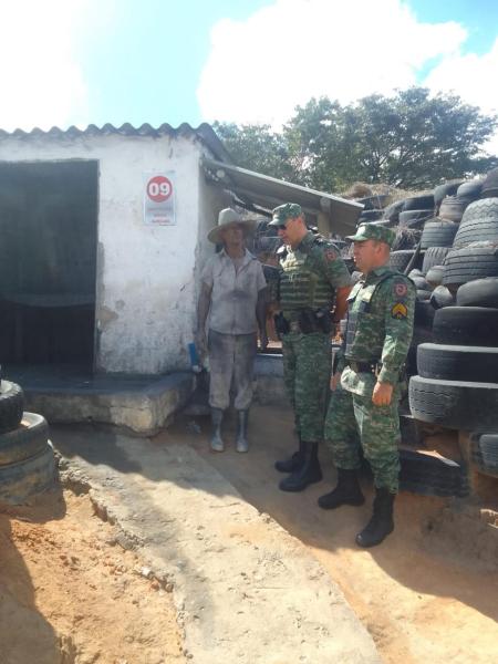 Homem com roupa suja e chapéu conversando com dois soldados em um local com pneus empilhados ao fundo.