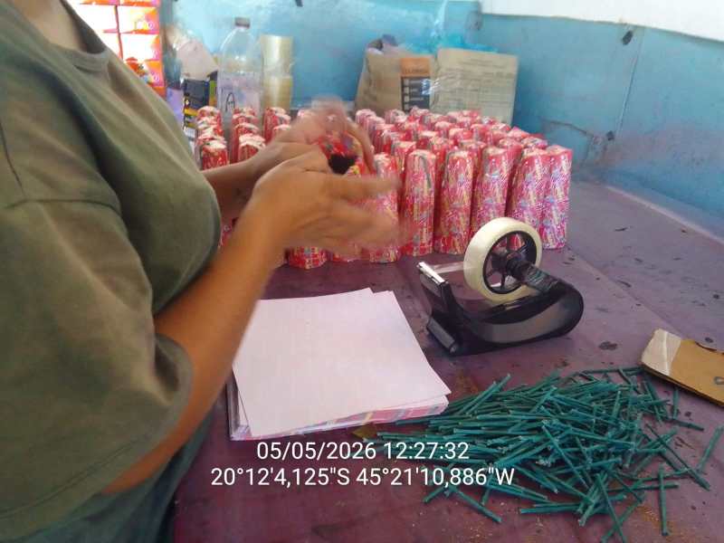 Pessoa organizando materiais de festa em uma mesa, com copos decorados e ferramentas de escritório ao fundo.