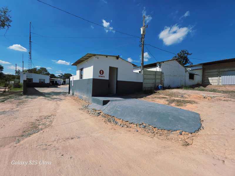 Imagem de uma construção branca com detalhes em cinza, situada em uma área rural. A estrutura apresenta uma rampa de acesso e está rodeada por um terreno de terra batida. No fundo, há outras construções e um poste de eletricidade sob um céu azul com algumas nuvens.