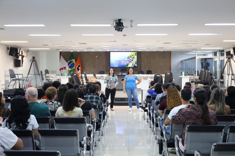 Duas mulheres em uma apresentação frente a um público em um auditório. Ao fundo, uma tela com uma imagem de natureza e bandeiras brasileiras.