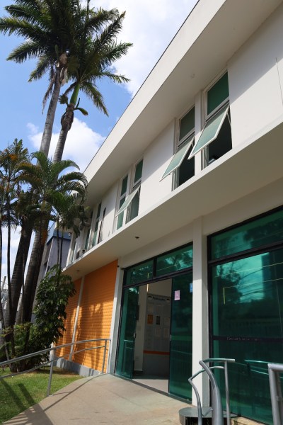 Fachada de um edifício moderno com janelas verdes e entrada de vidro, cercado por palmeiras e vegetação.