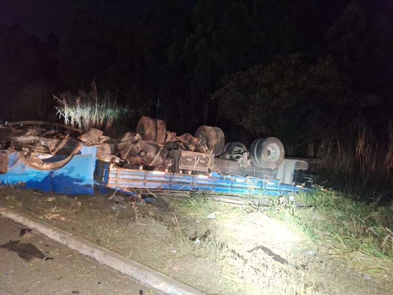 Caminhão tombado à noite em área de vegetação, mostrando a estrutura danificada e as rodas para cima.