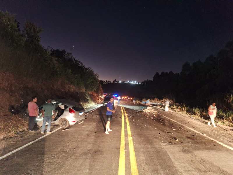 Cena de um acidente de carro à noite em uma estrada, com vários indivíduos próximos a veículos danificados e detritos espalhados na pista.