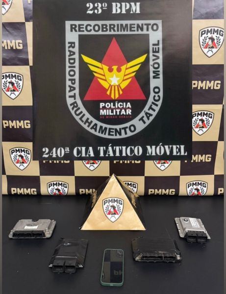 Imagem mostrando objetos apreendidos, incluindo eletrônicos, dispostos em uma mesa com um fundo que exibe o emblema da Polícia Militar de Minas Gerais.