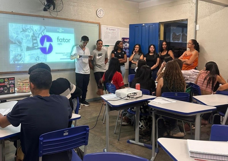 Imagem de um workshop em sala de aula, com um grupo de jovens apresentando enquanto outros alunos assistem. Um projetor exibe o título 'Workshop - Definições' na parede.