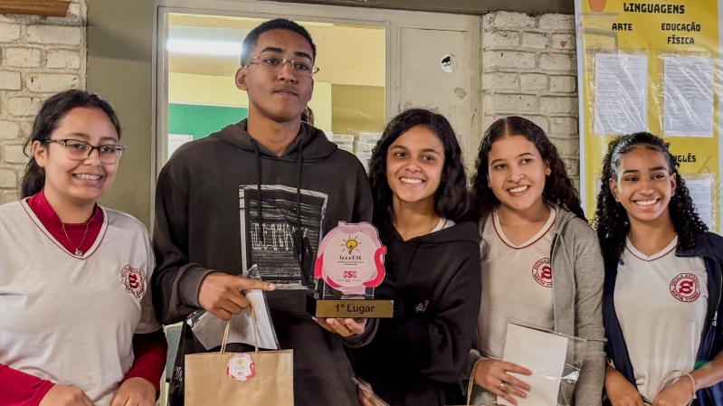 Grupo de estudantes sorrindo em ambiente escolar, segurando um prêmio de '1º Lugar'.