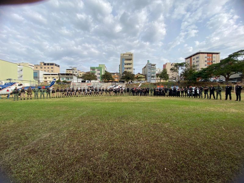 Grupo de policiais em formação em um campo, com prédios e um helicóptero ao fundo sob um céu nublado.