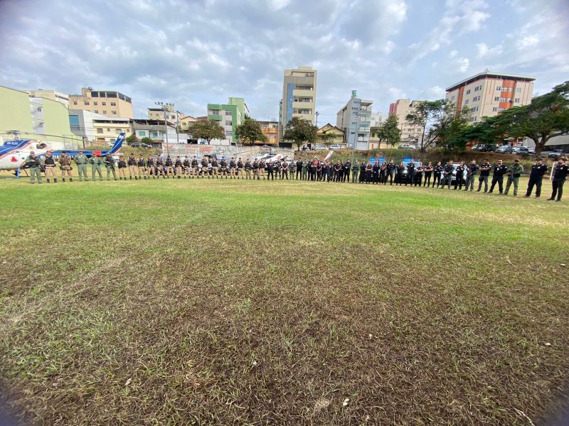 Uniformizados em uma linha, policiais e membros de equipe de segurança se organizam em um campo, com prédios ao fundo e um helicóptero próximo.
