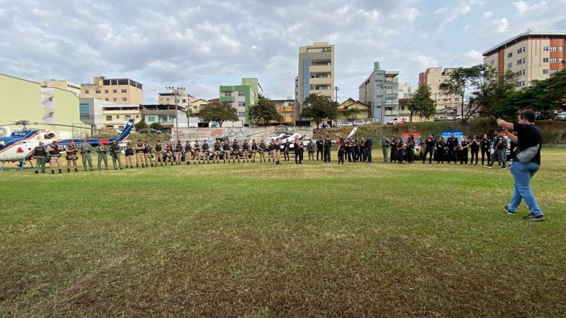 Grupo de policiais e agentes de segurança em pé em uma área aberta, com um helicóptero ao fundo, sob um céu nublado e edifícios urbanos.