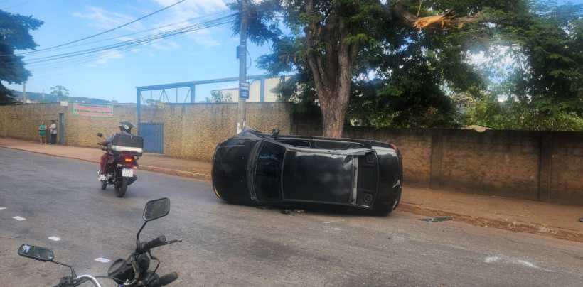 Carro preto capotado na rua, com motocicleta passando ao lado.