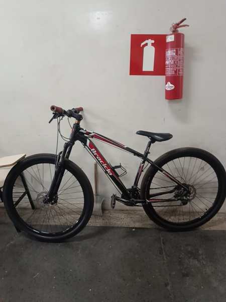 Uma bicicleta mountain bike preta com detalhes vermelhos encostada em uma parede, ao lado de um extintor de incêndio.
