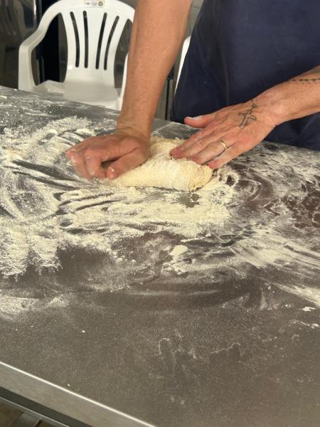 Mãos amassando massa de farinha em uma superfície de trabalho, com uma cadeira ao fundo.