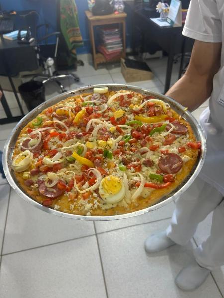 Uma pizza grande e colorida, coberta com linguiça, presunto, pimentões, cebolas, tomate, azeitonas e ovos cozidos, sendo segurada por uma pessoa.