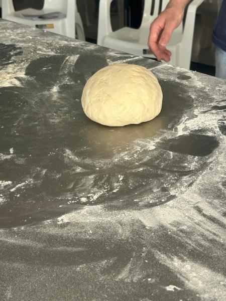 Massa de pão em formato redondo sobre uma superfície enfarinhada, com uma mão visível ao fundo.
