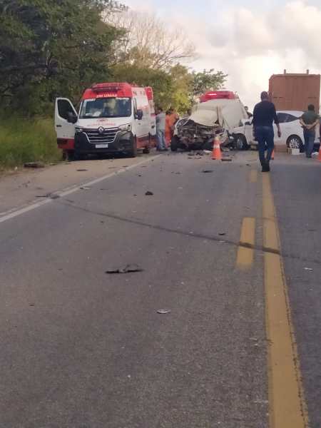 Cena de um acidente de trânsito, com duas ambulâncias paradas e um veículo danificado na estrada. Há homens conversando e destroços espalhados pelo chão.