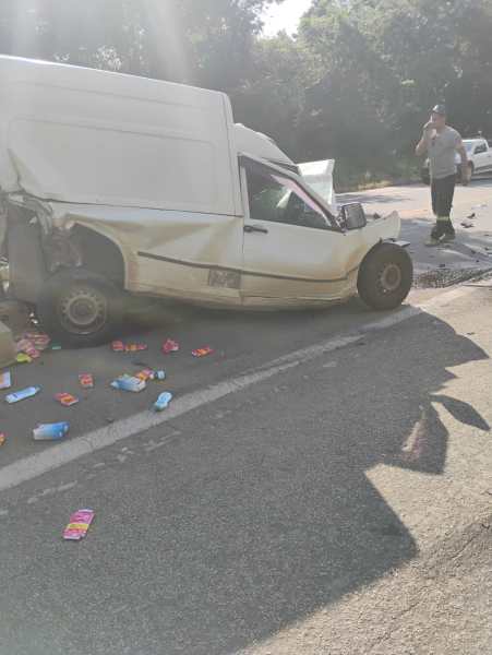 Um acidente de carro envolvendo uma van branca danificada, com parte de sua carroceria amassada. Vários produtos estão espalhados ao redor da van na estrada. Um homem está de pé próximo ao veículo, possivelmente avaliando a situação.