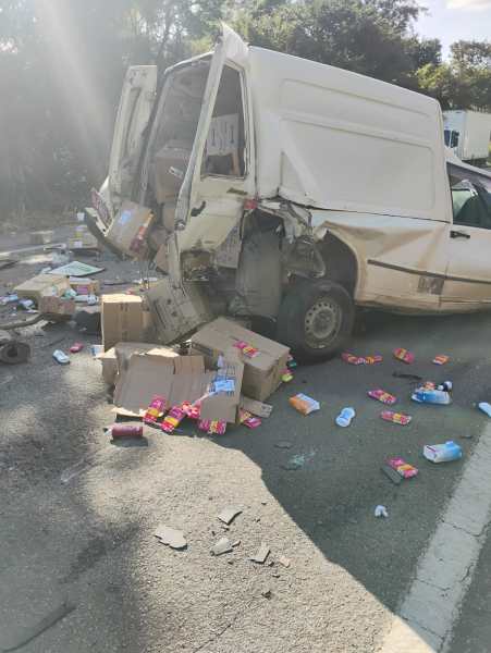 Furgão acidentado na estrada, com a traseira danificada e caixas de produtos espalhadas ao redor.
