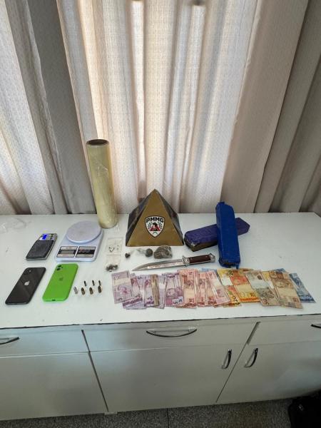 Mesa com objetos diversos, incluindo telefones celulares, um peso, munições, dinheiro em notas e uma base triangular com logotipo.
